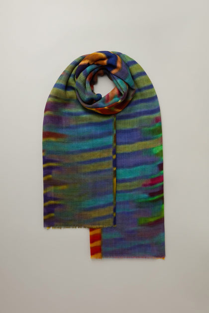 The Brushstroke Stripe Wool Blend Scarf, with vibrant multicolored stripes in blue, green, yellow, and red, is showcased against a neutral background. Perfect for versatile styling, it offers a lively enhancement to any outfit.