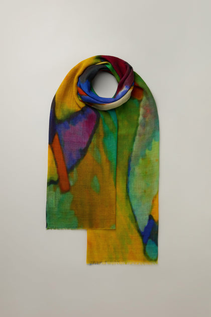 The Painterly Wool Blend Scarf features a vibrant abstract print in yellow, green, blue, and red shades. It's versatile for styling and is elegantly rolled at one end against a plain light background.