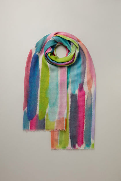 The Shaded Watercolour Stripe Wool Blend Scarf showcases vertical watercolor-style stripes in pink, blue, green, and purple on a light background.