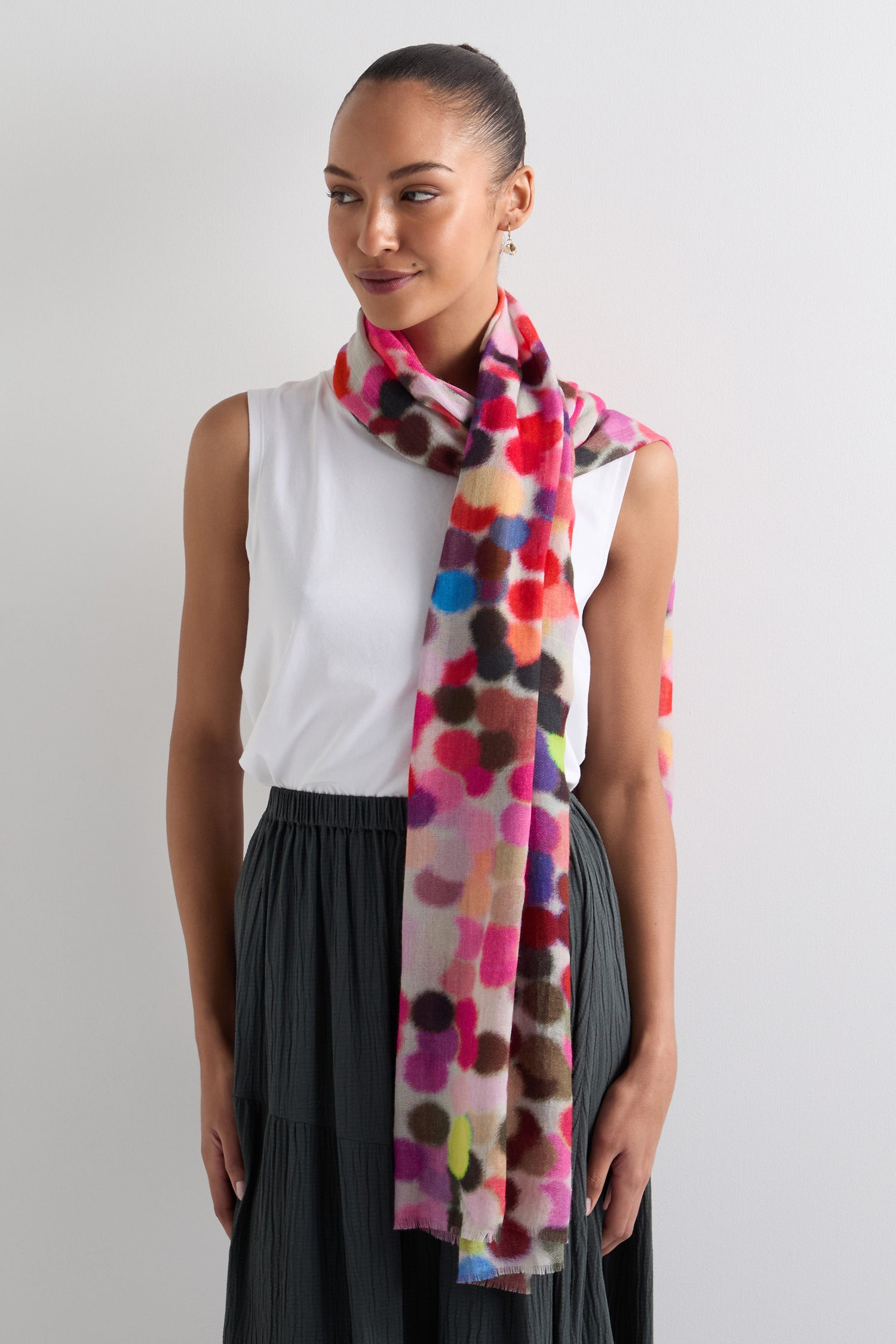 A person in a sleeveless white top and dark skirt stands against a plain background, elegantly draped with the Dotty About You Wool Blend Scarf. The colorful polka-dot pattern adds a playful touch to their ensemble.