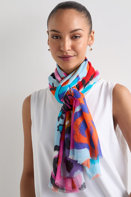 The person is wearing a vibrant Painted Stars Cotton Blend Scarf over a white top, smiling slightly against a neutral background.