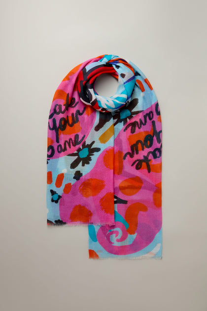 The Painted Stars Cotton Blend Scarf features an artistic star motif and abstract patterns in pink, blue, orange, and black. Displayed flat against a plain background, this colorful accessory adds a whimsical touch to any outfit.