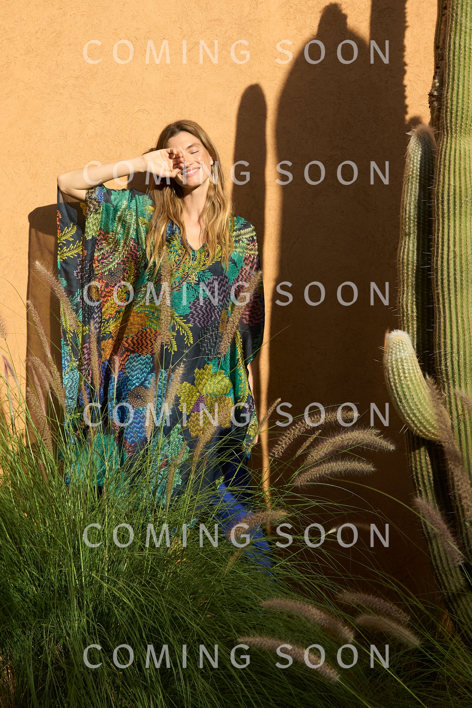 A woman wearing the Pixelated Petals Silk V-Neck Kaftan with a pixel floral print stands outside by cacti and tall grass, shading her eyes from the sun. "COMING SOON" is displayed over the image.