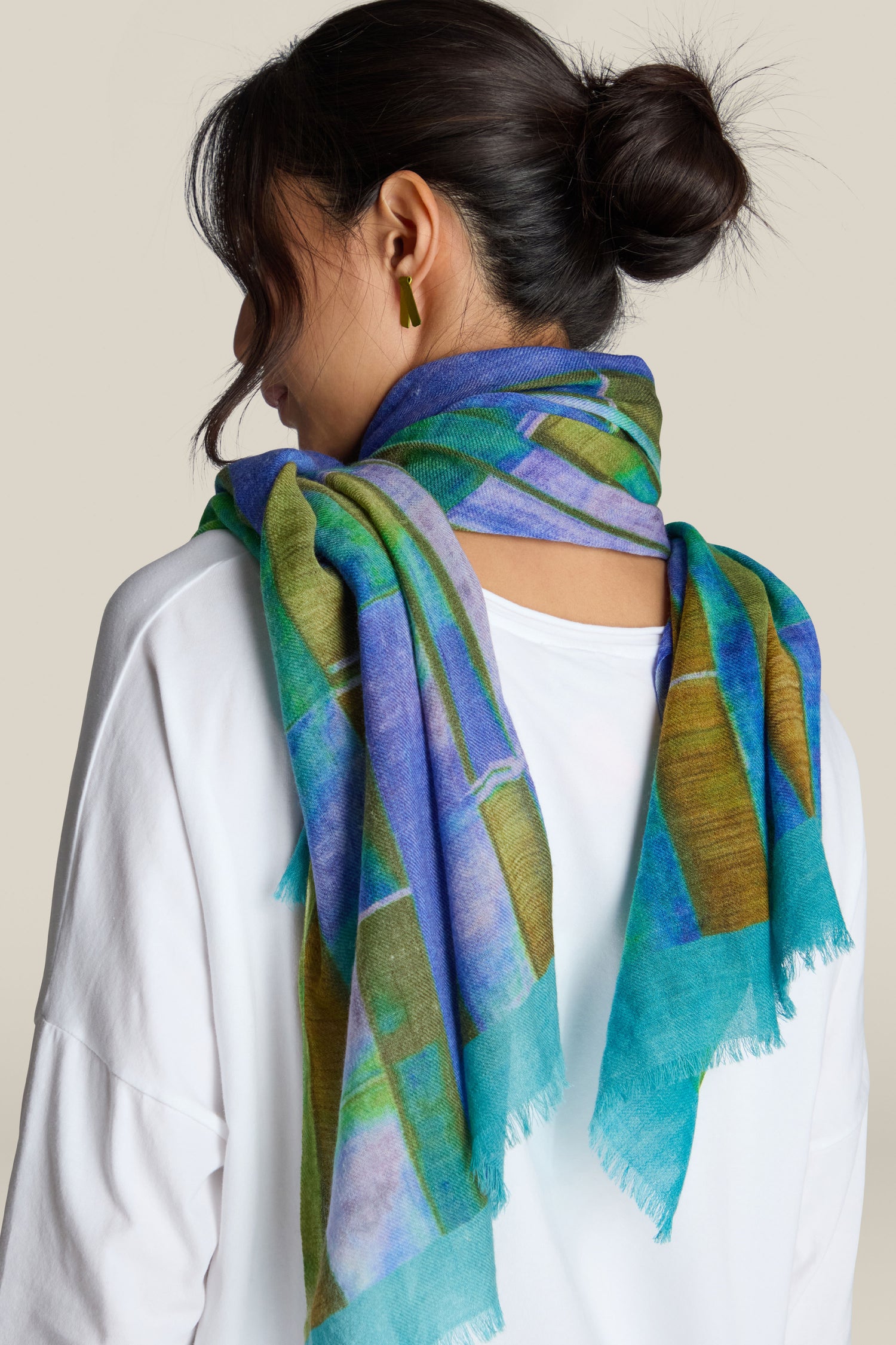 A person with dark hair in a bun is wearing a white top and a vibrant Fragment Wool Scarf featuring blue, green, and yellow patterns. Made in India from premium wool, the scarf adds an eye-catching touch as the person is shown from the back.