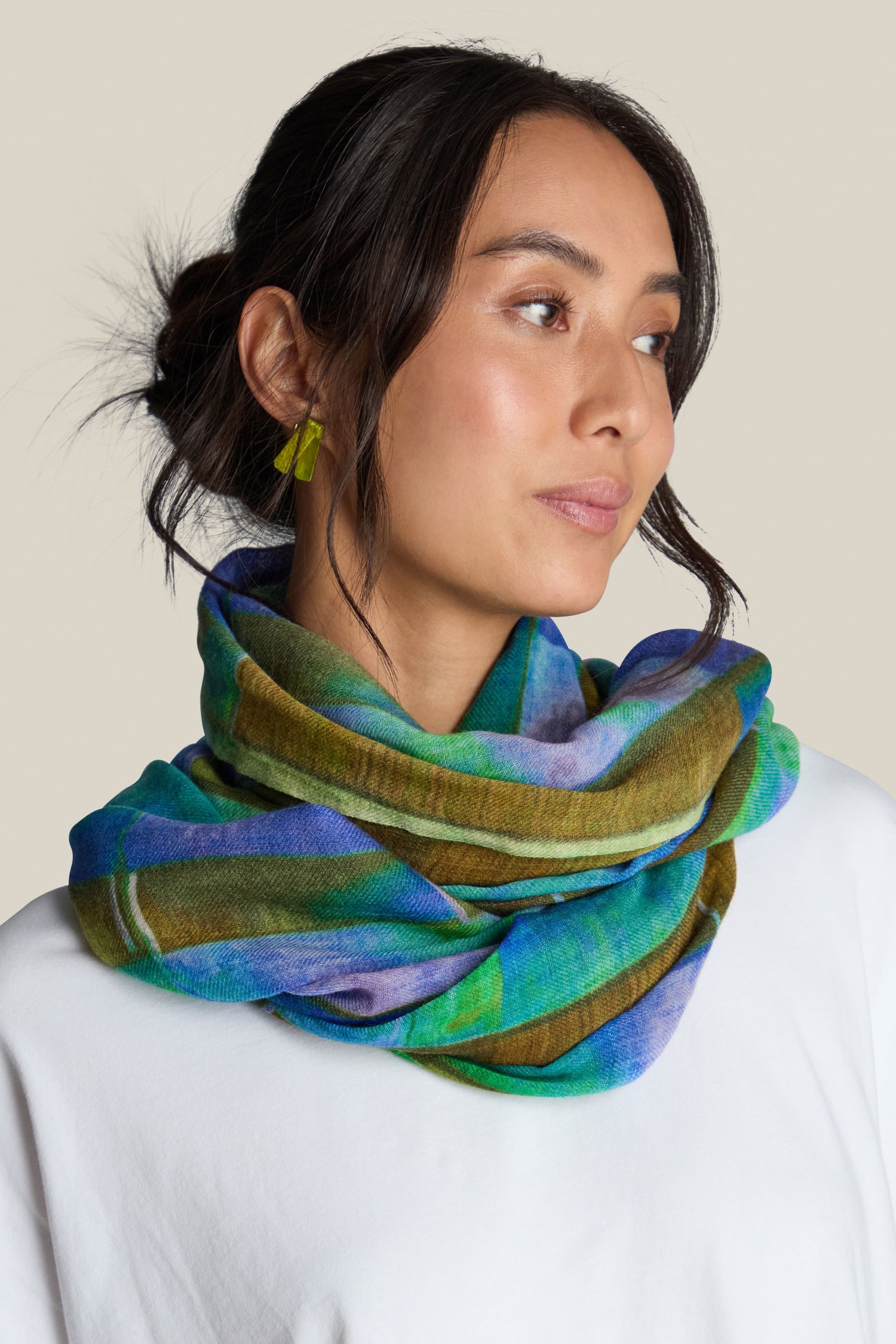 A person with dark hair tied back wears green geometric earrings and a colorful, multi-patterned Fragment Wool Scarf made in India. They are looking to the side and wearing a white top.