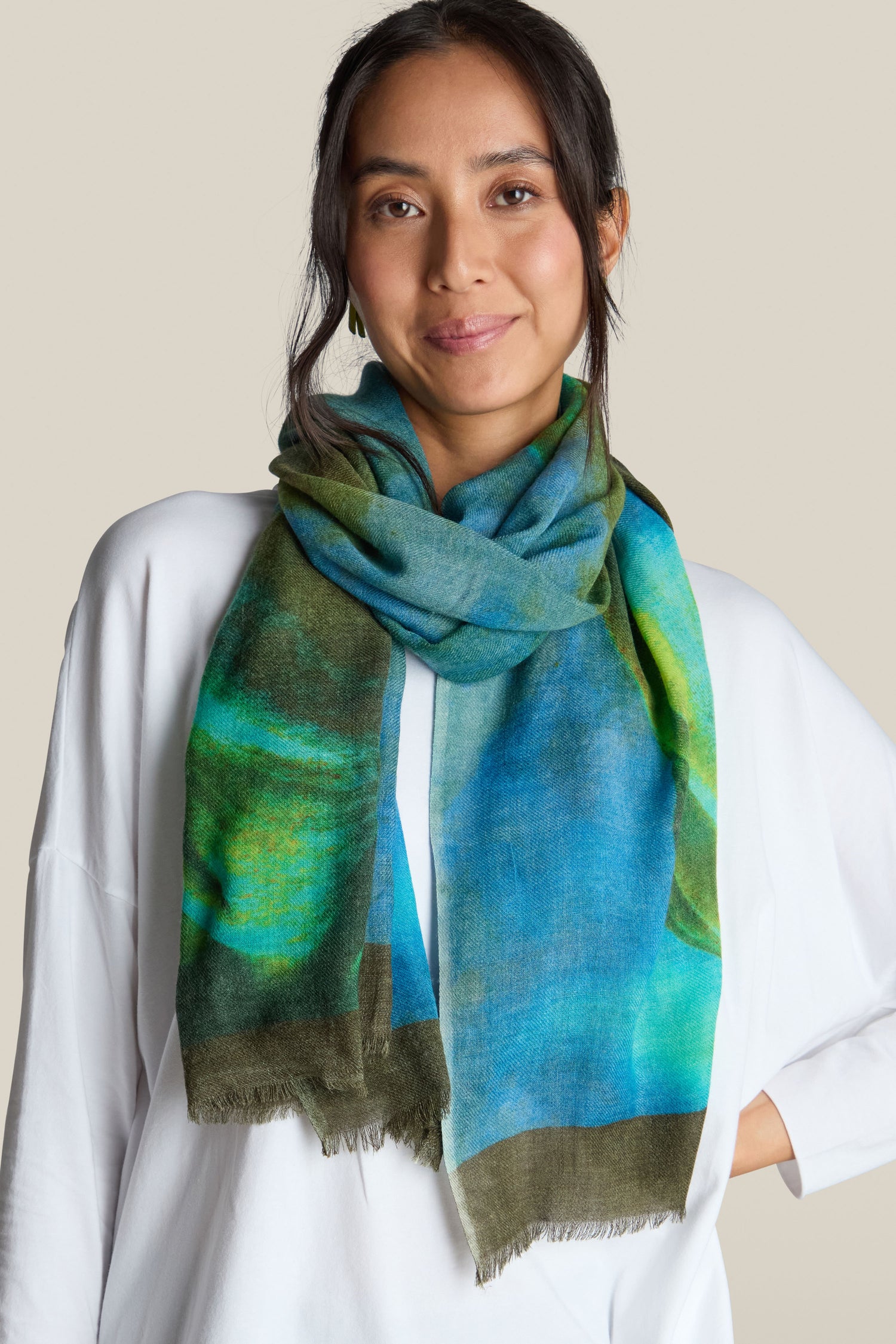 A person with dark hair is wearing a white top and the Ocean Deep Wool Scarf in sea-tone colours, standing against a plain beige background.