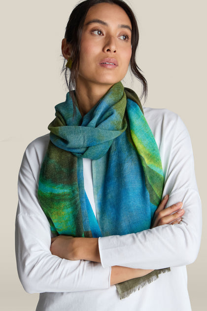 A person with long dark hair is wearing a white top and a vibrant green and blue Ocean Deep Wool Scarf. They are standing with their arms crossed and looking to the side.