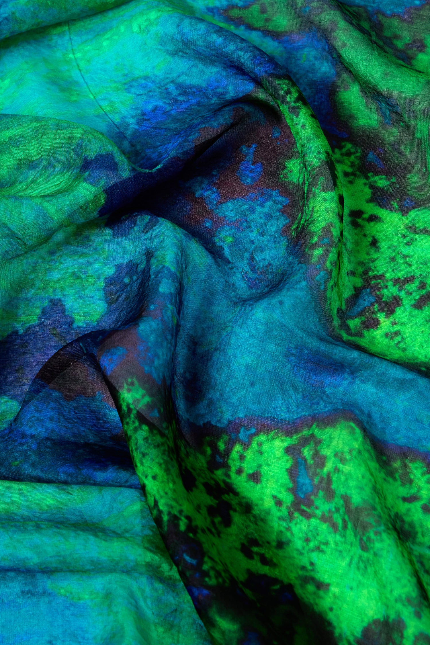 A close-up image of a fabric with vibrant blue and green tones, resembling the Galaxy Silk Scarf, showcases a mesmerizing tie-dye pattern and textured surface.