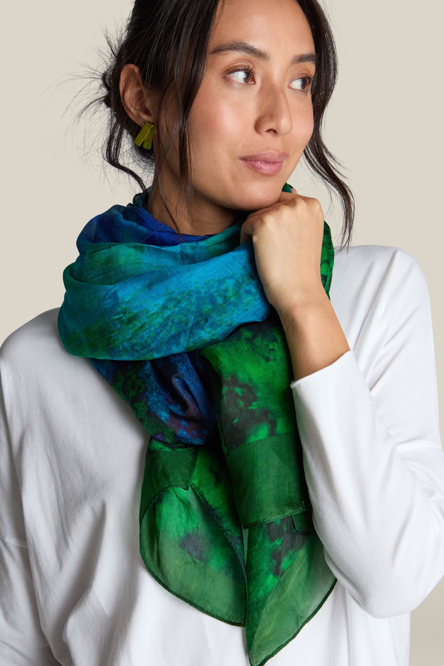 A person wears a white top paired with the luxurious Galaxy Silk Scarf in a green and blue pattern, looking to the right with a neutral expression.