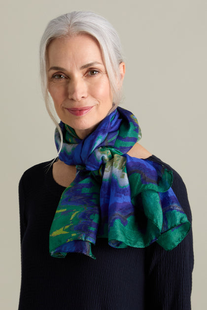 An older woman with long, gray hair tied back, wearing a dark sweater and a vibrant blue and green Mineral Silk Scarf around her neck, smiles slightly against a neutral background.