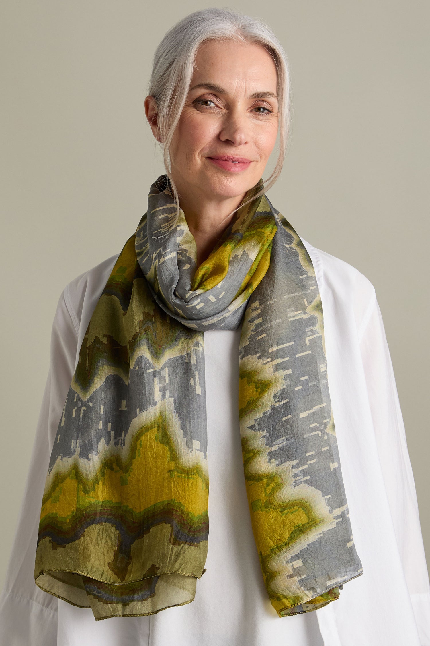 An older woman with gray hair, wearing a white top and a luxurious Mineral Silk Scarf, smiles gently at the camera.