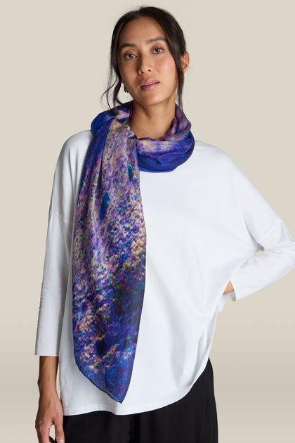 A person with a relaxed expression wears a white top and the Quartz Silk Scarf, showcasing vibrant purple and blue quartz crystal formations, standing against a plain background.