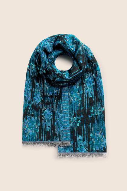 A folded Midnight Stitch Scarf, featuring an elegant combination of blue and black adorned with intricate floral patterns and fringed edges, is beautifully displayed against a beige background.