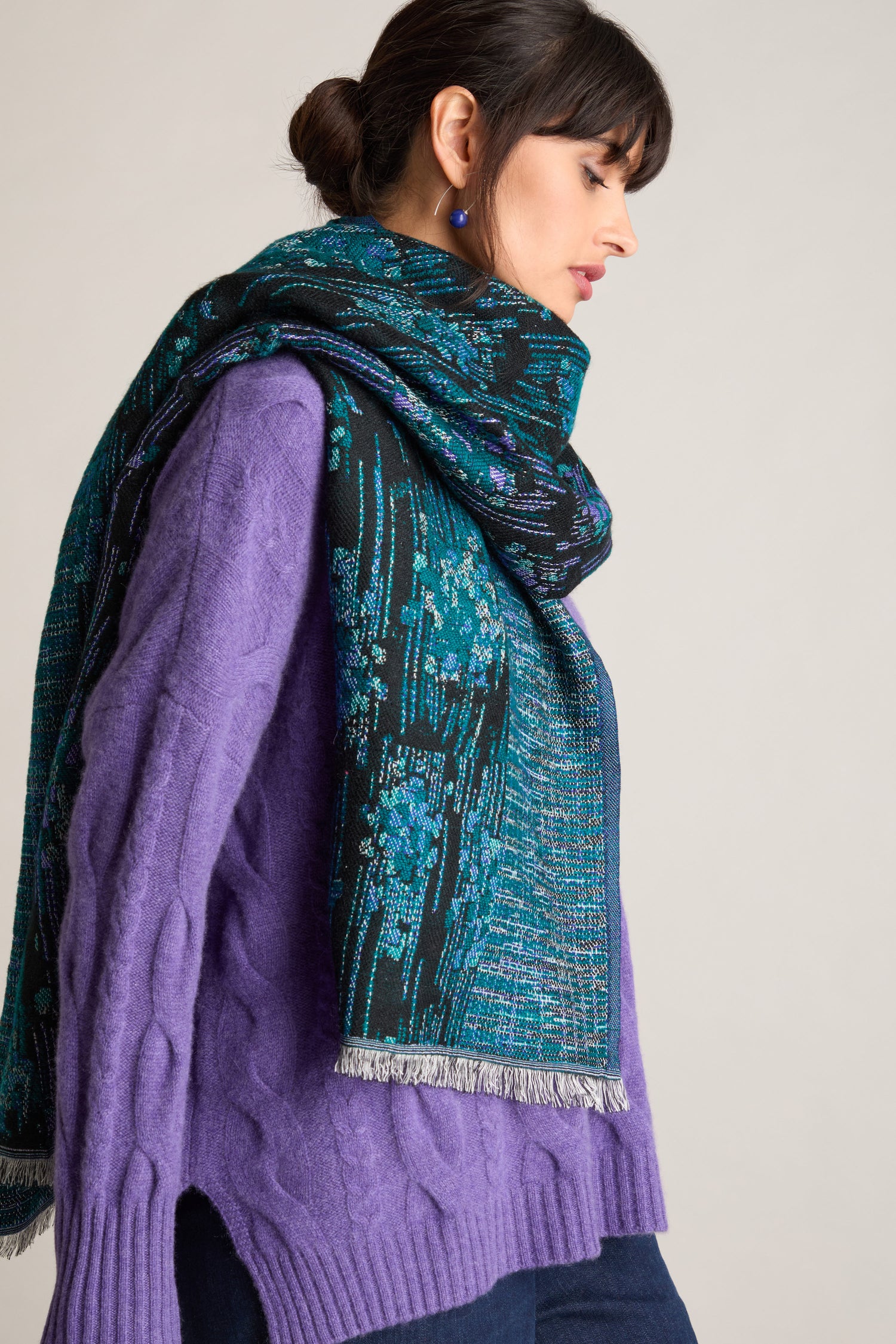 A person with dark hair tied back and facing sideways is wearing a luxurious Midnight Stitch Scarf. The scarf, draped over their shoulders, features intricate patterns in black and teal. They are dressed in a purple sweater and dark blue jeans.