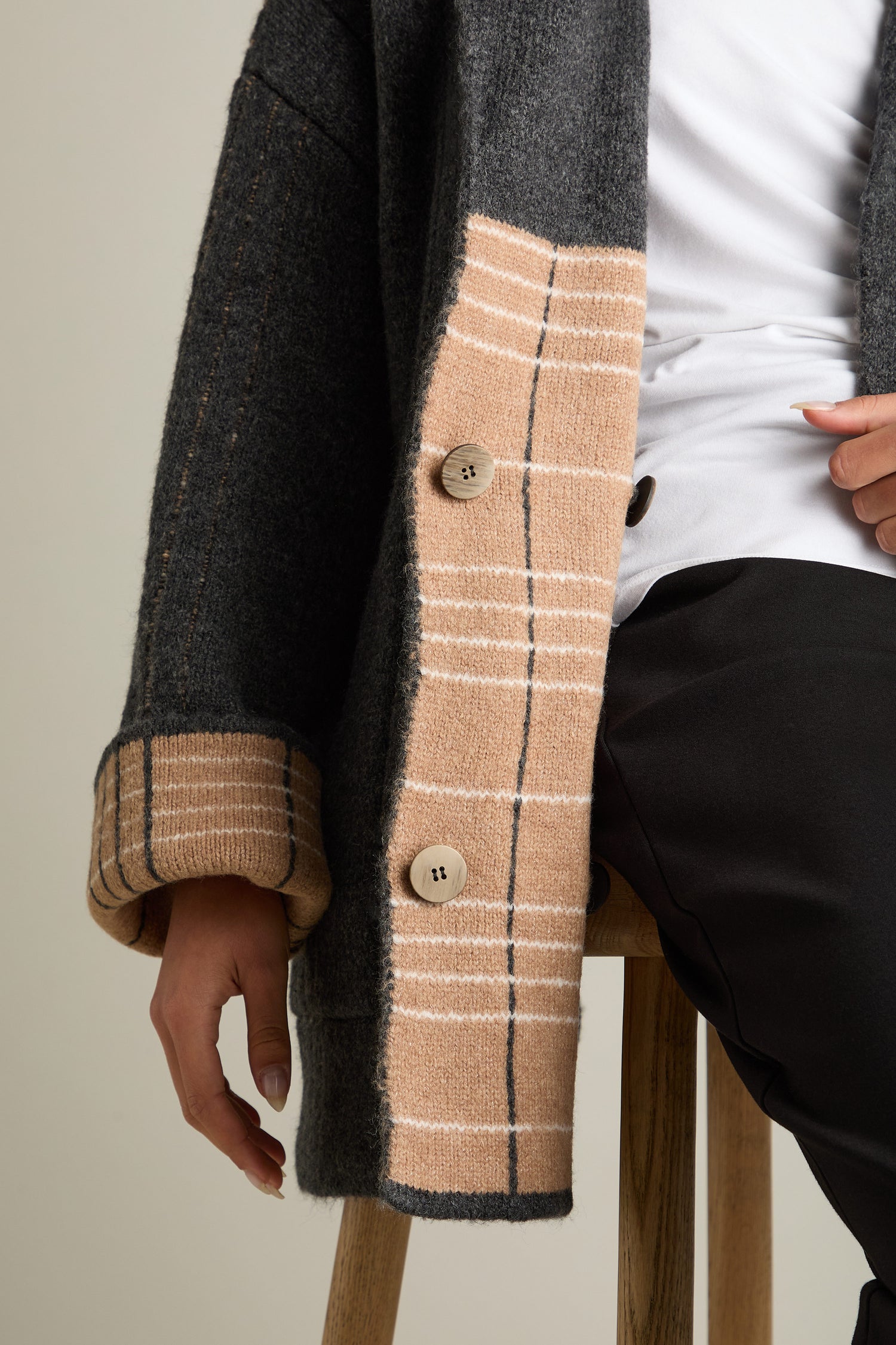 A person sitting on a stool is wearing the Check Detail Jacket, featuring a dark wool blend with check detail and large buttons, stylishly layered over a white top.