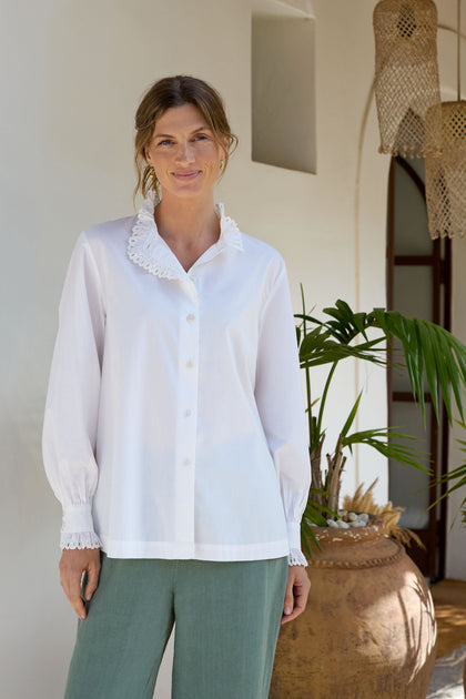 A woman stands indoors, smiling, wearing a "Frill Collar Shirt" with delicate lace trim and cutwork embroidery, paired with green pants. She stands near a decorative clay pot and plants.