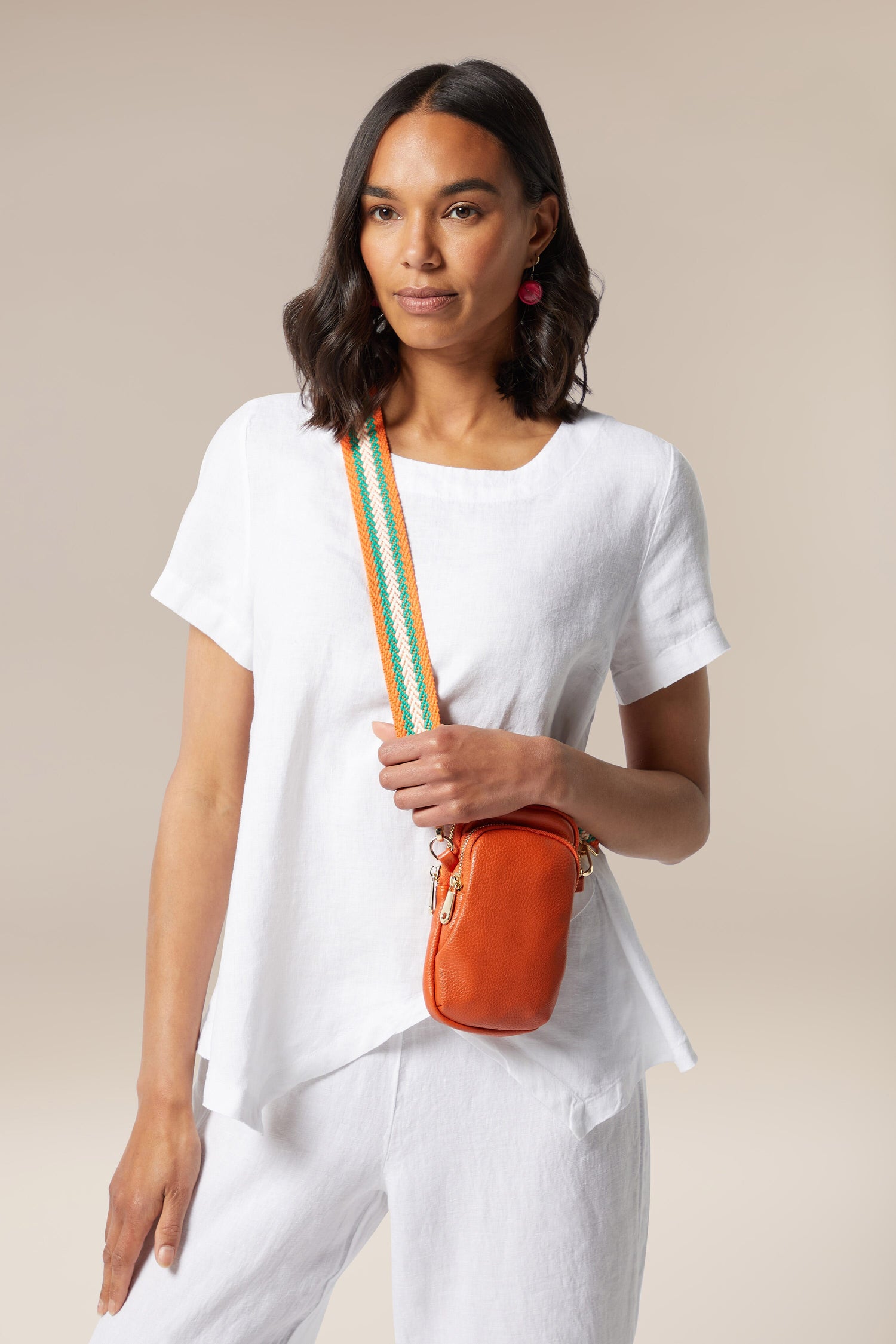 Kris-Ana, a woman wearing white pants, showcases her style-led designs through her vibrant orange Mini Crossbody Bag.