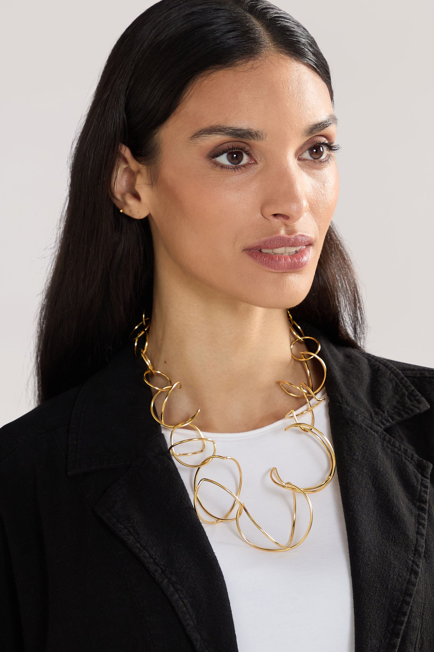 A person with long, dark hair wears a contemporary Handmade Links Necklace crafted from 24k gold plated brass and a black jacket over a white top, looking to the side against a neutral background.