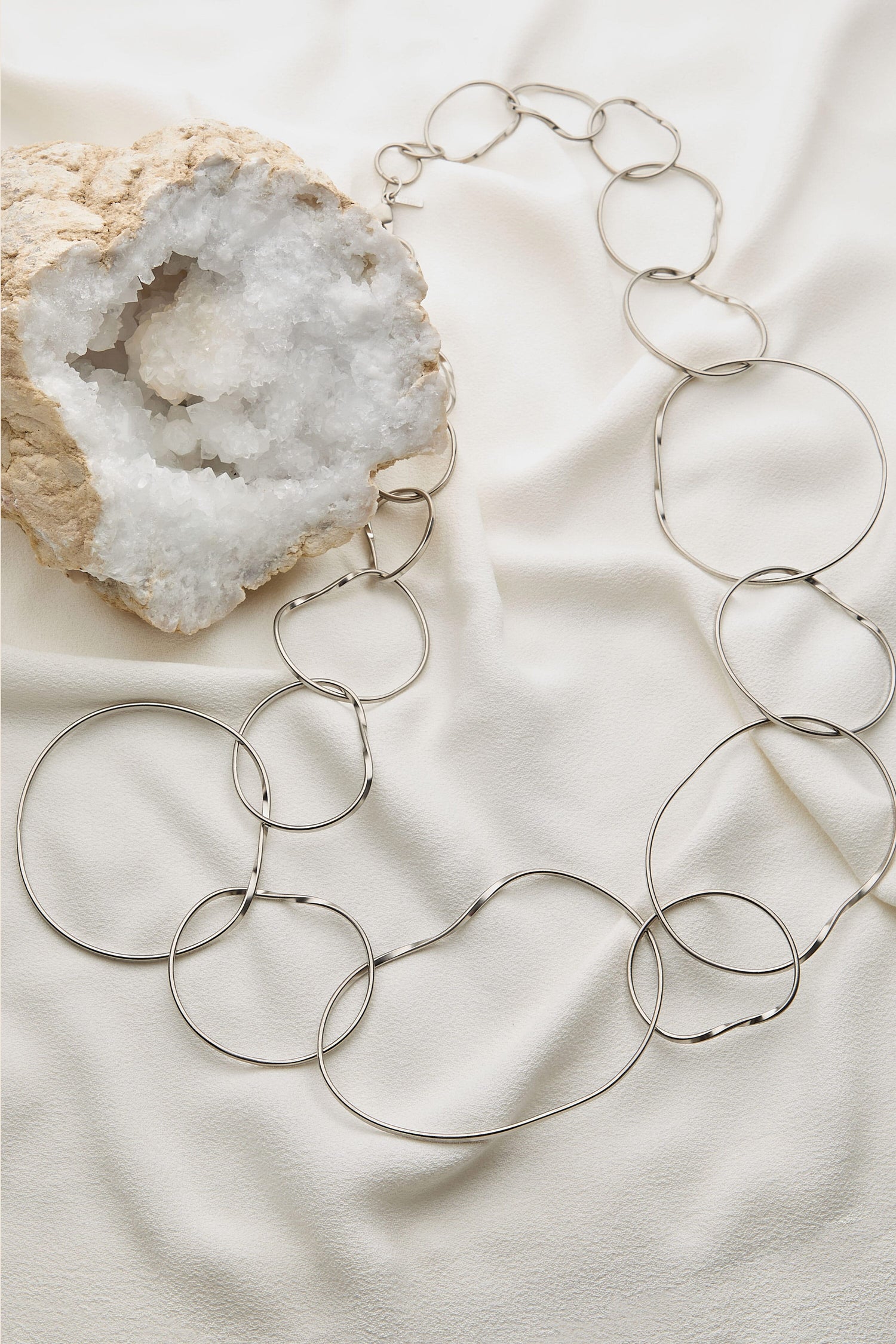 The Handmade Silver Hoops Necklace, featuring meticulously crafted circular loops of various sizes by a renowned Greek designer, is elegantly draped over a light-colored fabric next to a partially opened geode. This exquisite piece perfectly encapsulates the essence of handmade jewelry with its intricate design and artisanal charm.