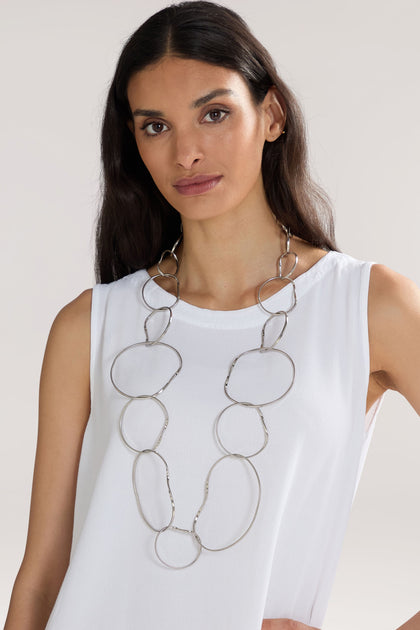 A woman with long dark hair wears a white sleeveless top and a long, chunky Handmade Silver Hoops Necklace by a renowned Greek designer. She looks directly at the camera against a neutral background.