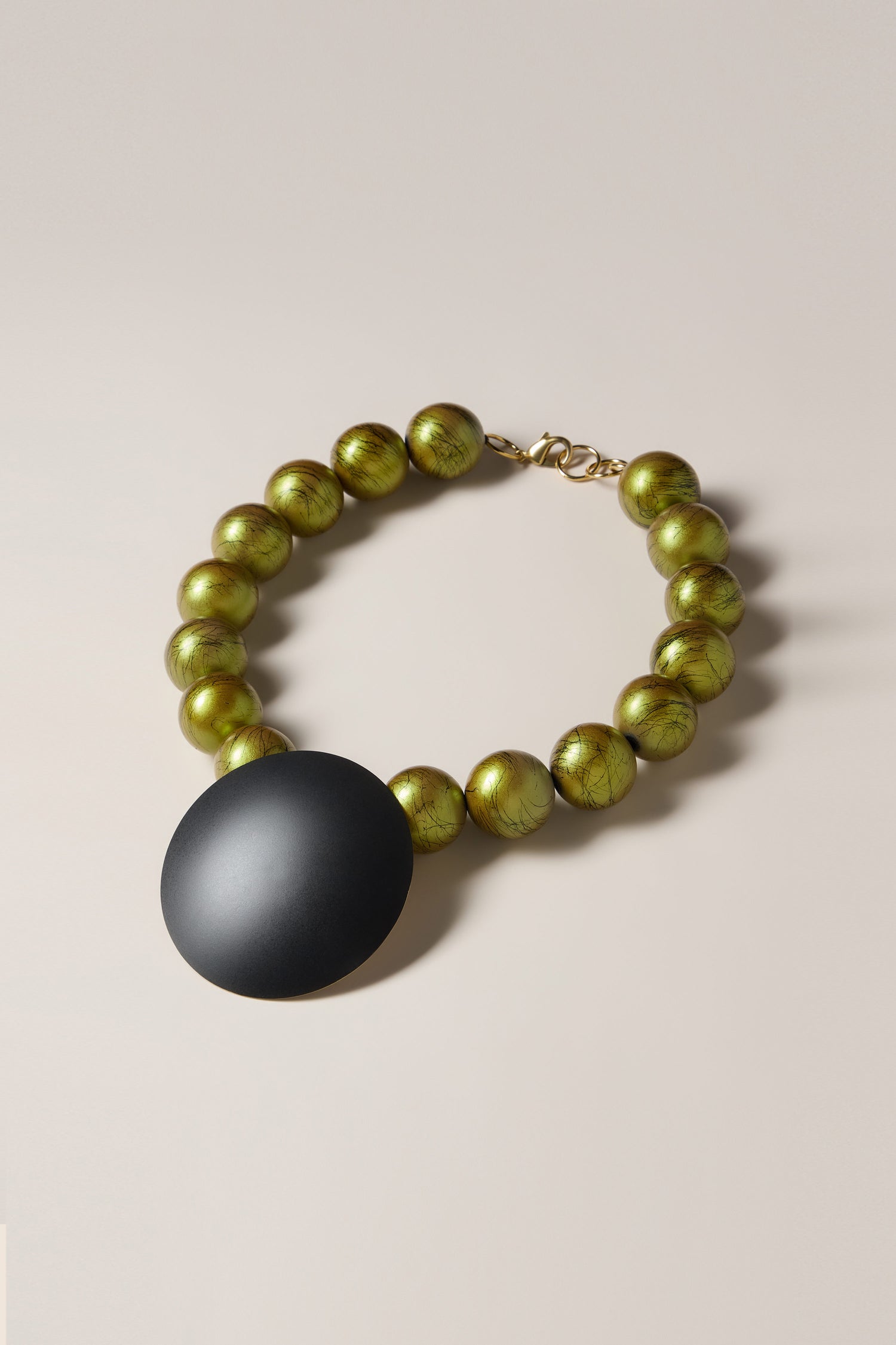 The Handmade Eclipse Necklace is a statement piece, featuring a striking black circular pendant with a clasp, encircled by vibrant green spherical beads on a light beige backdrop, creating a modern and stylish look.