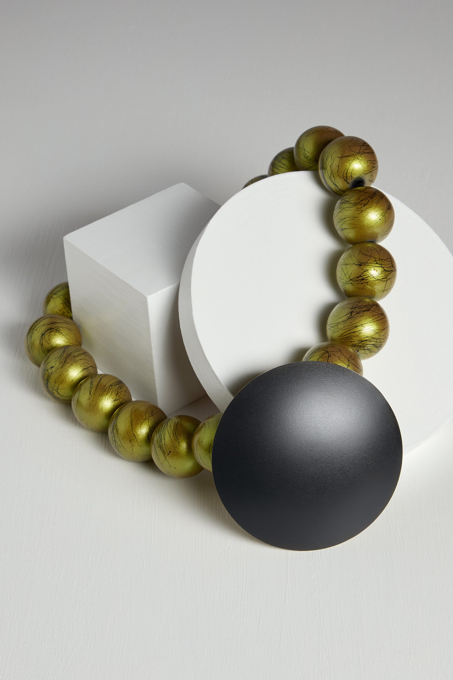 The Eclipse Short Necklace is an eye-catching piece of hand-crafted jewellery, featuring large round beads in a greenish-gold hue and a striking black circular pendant. Showcased on white geometric shapes, this exquisite necklace highlights the distinctive craftsmanship of a Greek designer.