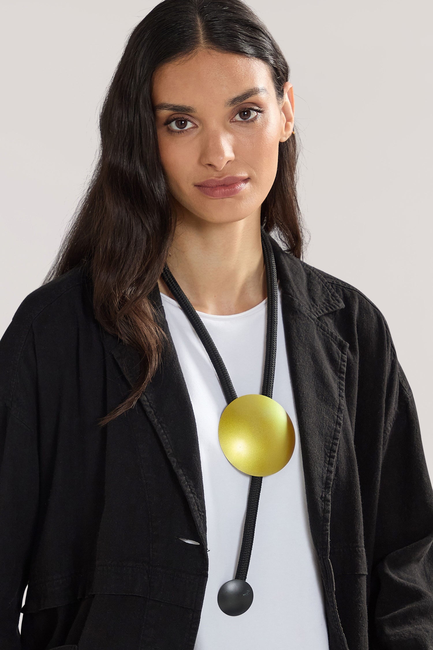 A person with long dark hair is wearing a white shirt, a black jacket, and the Handmade Aphelion Necklace, a large round yellow pendant on a black cord neckpiece, exuding a bold contemporary look reminiscent of pieces by a Greek jewellery designer.