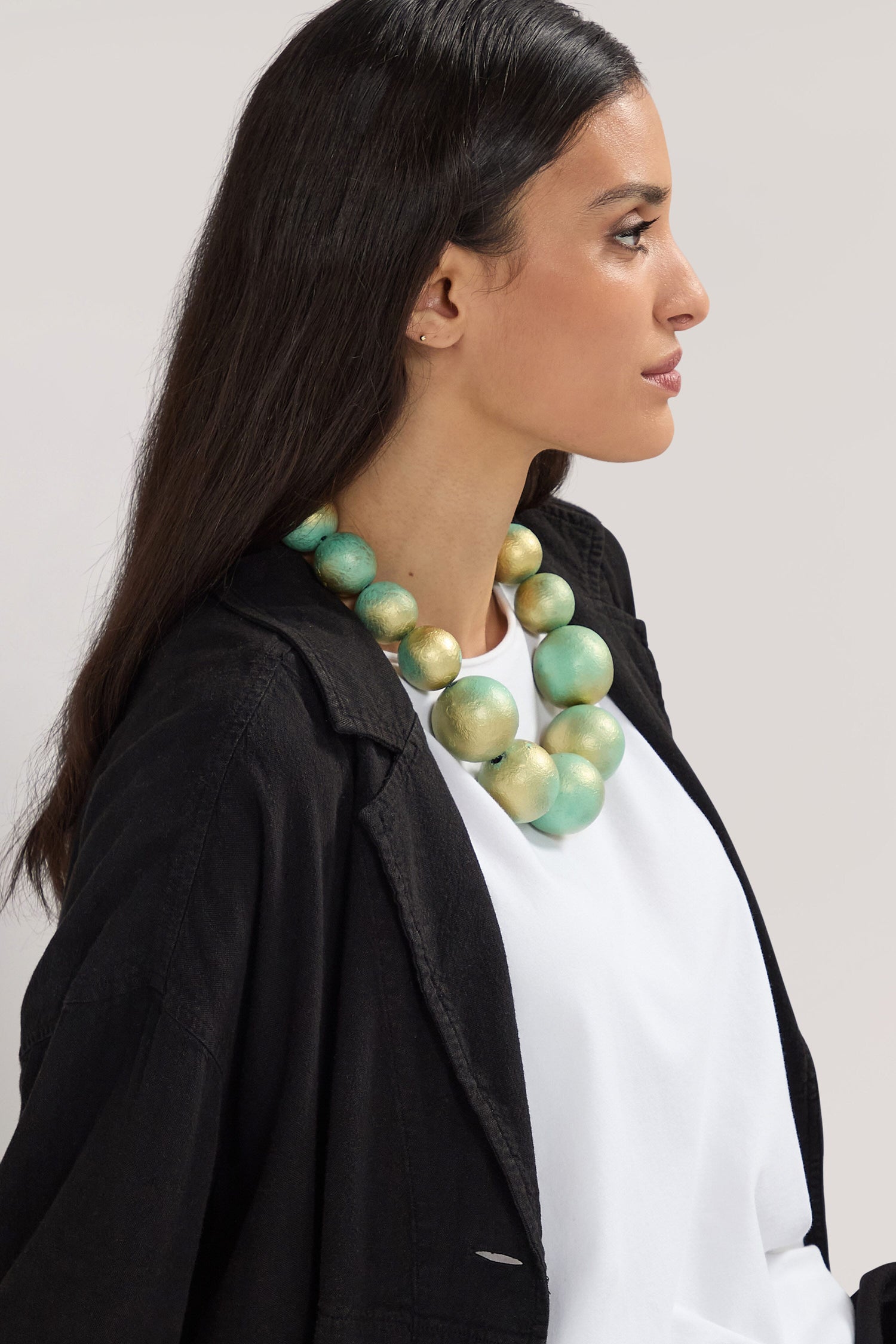 A person with long dark hair is wearing a white shirt, black jacket, and a Handmade Short Painted Spheres Necklace adorned with large green and gold beads from a Greek designer, looking to the side.