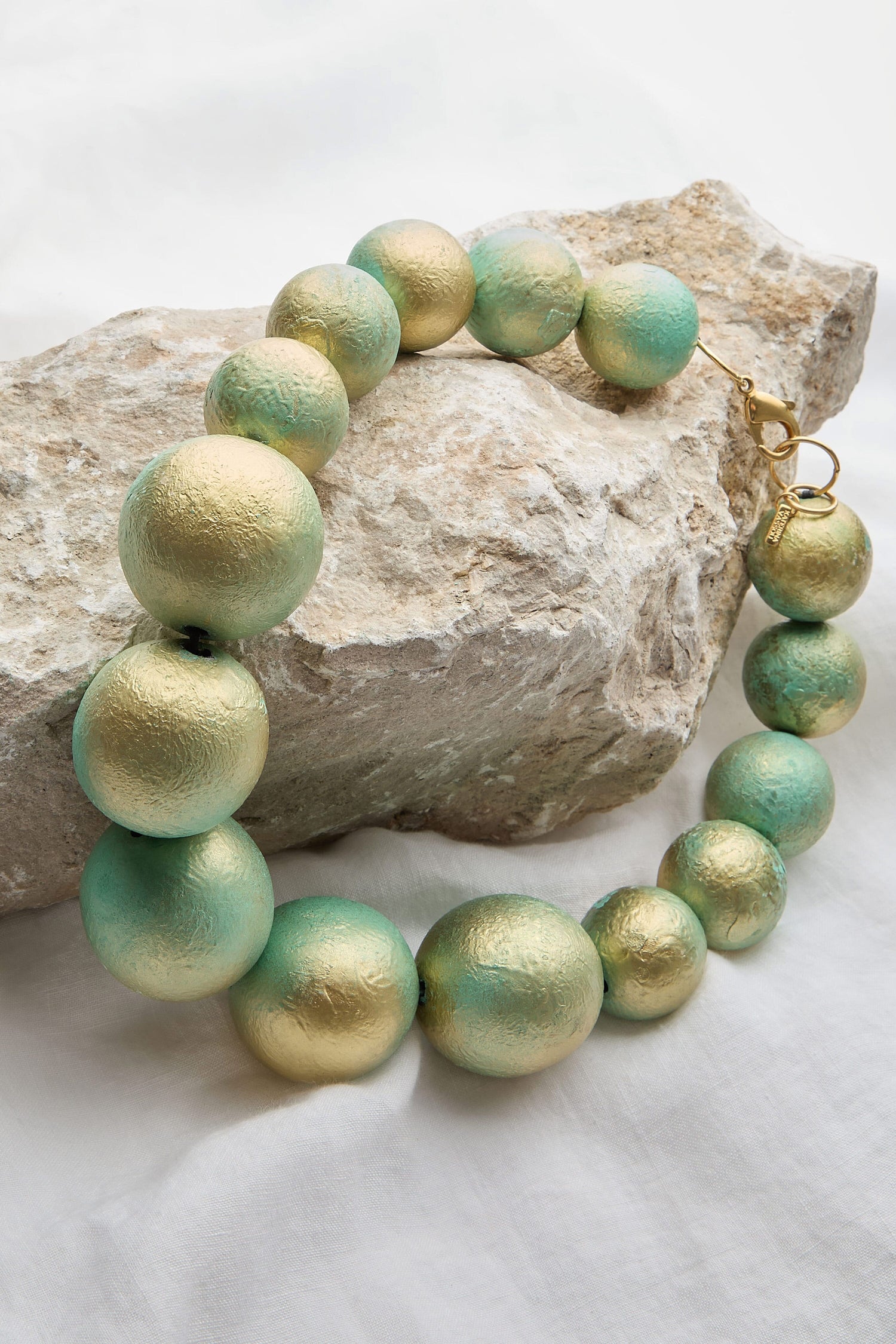 The Handmade Short Painted Spheres Necklace, featuring alternating large and small gold and green spherical beads crafted by a talented Greek designer, is displayed draped over a rough stone on a light fabric background.