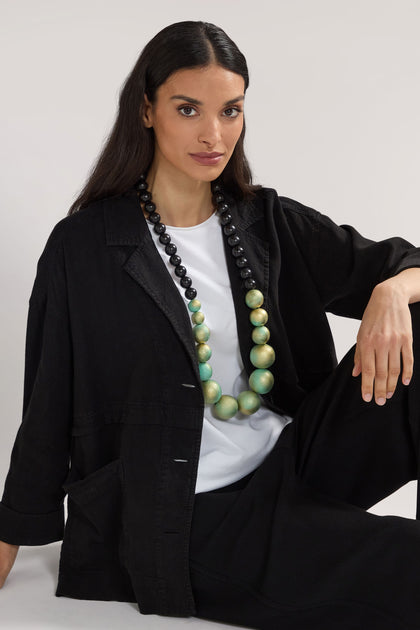 A seated person with long dark hair wears a black jacket over a white shirt, adorned with a bold Handmade Painted Spheres Necklace designed by a renowned Greek jewellery designer.