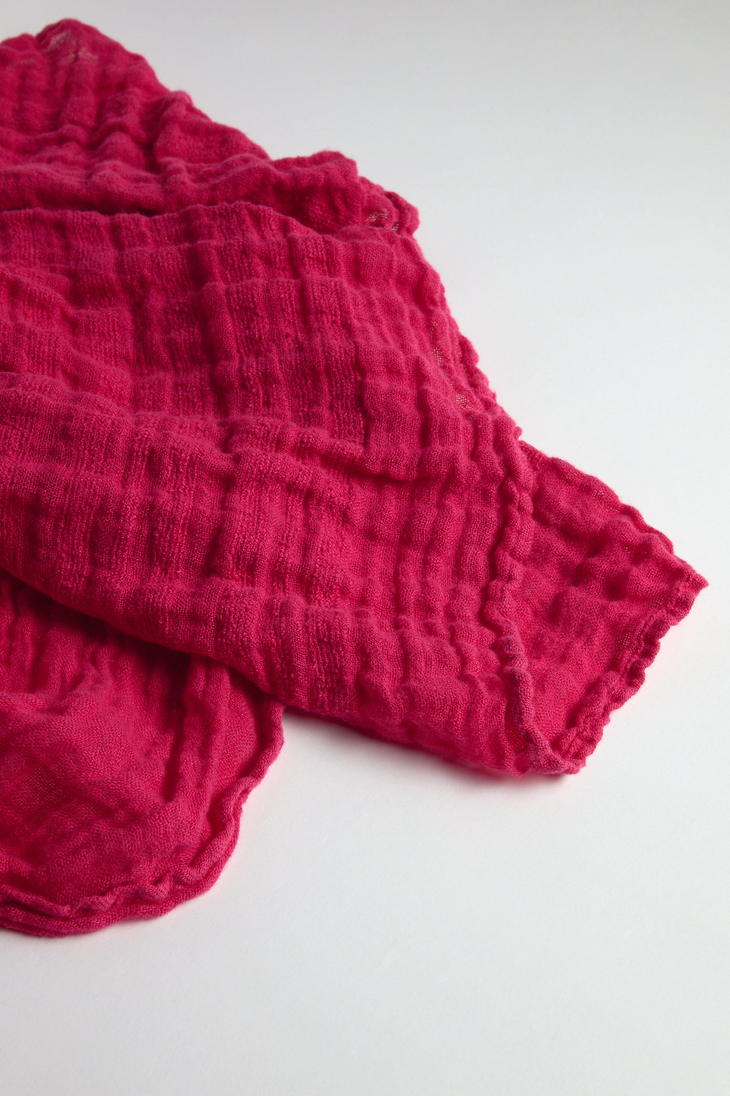 The Crinkle Linen Scarf, in bright pink, is gracefully displayed on a white surface, highlighting its textured design. This lightweight accessory adds vibrant color and style to any outfit, making it a versatile choice for all occasions.