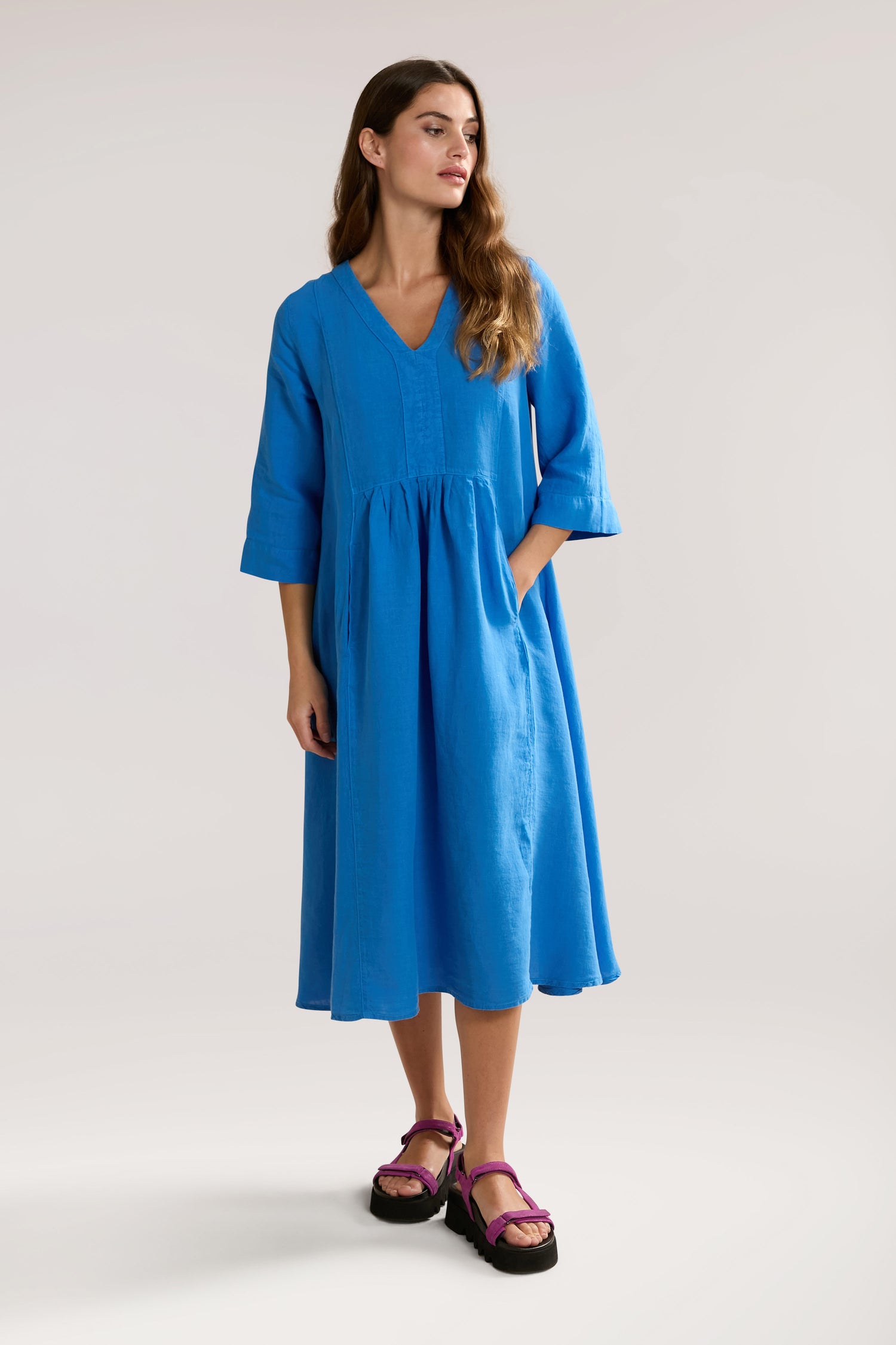 Woman wearing a loose-fitting, bright blue Organza Linen Pleat Front Dress with three-quarter sleeves and pink sandals stands against a plain background. She has long, wavy hair.