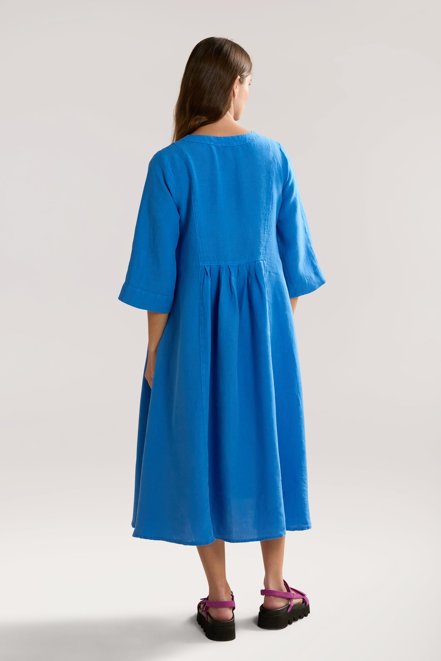 A person with long hair wearing a blue, knee-length Organza Linen Pleat Front Dress stands facing away, showcasing the back design with gathered pleats.