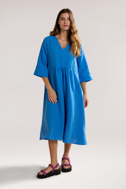 A woman is standing and wearing a bright blue, knee-length Organza Linen Pleat Front Dress with three-quarter sleeves and pink sandals. She has long, wavy brown hair and a neutral expression.