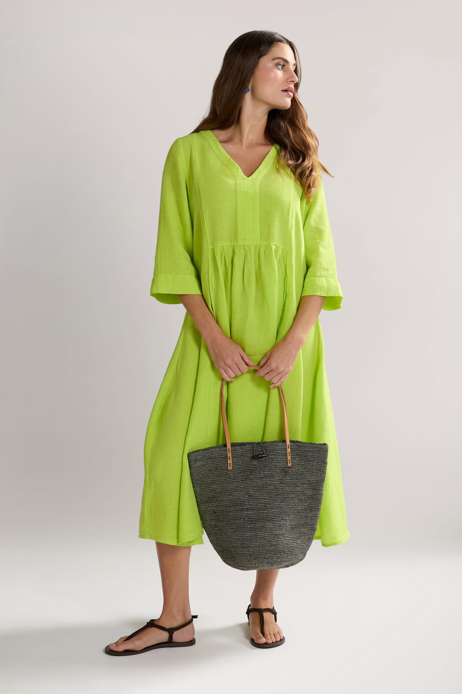 A woman in a bright green, Organza Linen Pleat Front Dress holds a large gray tote bag and stands against a plain background, wearing black sandals and looking to her left.