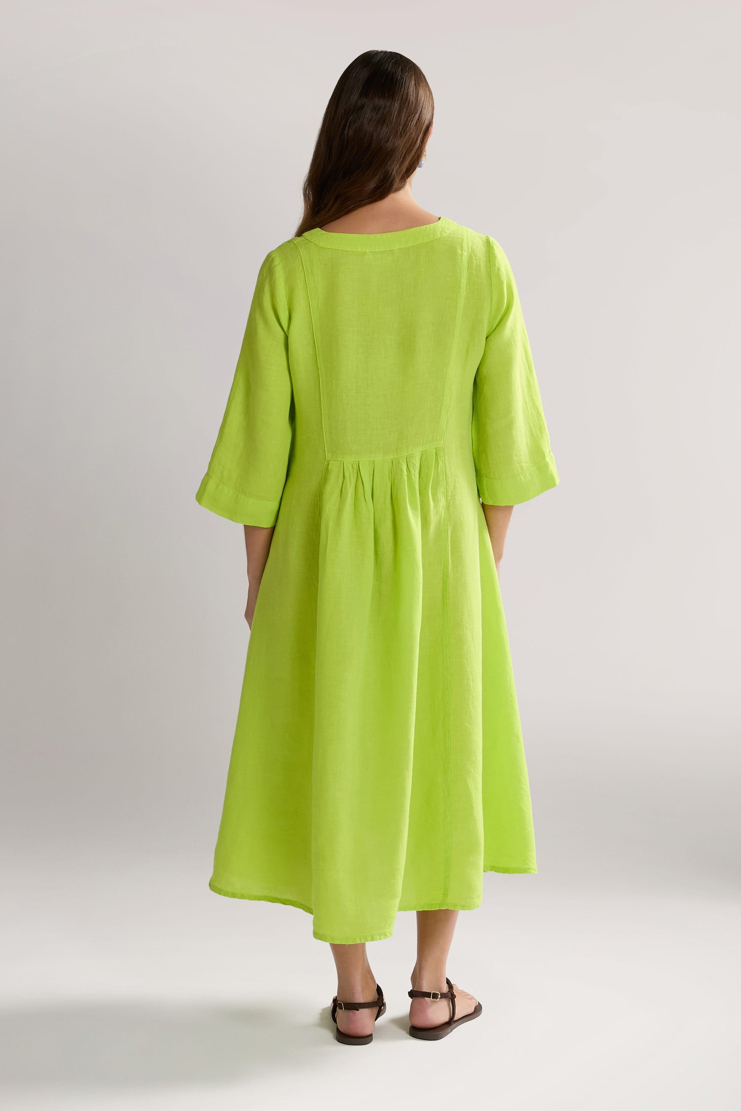 A person with long hair stands facing away, wearing a bright green, Organza Linen Pleat Front Dress and brown sandals.