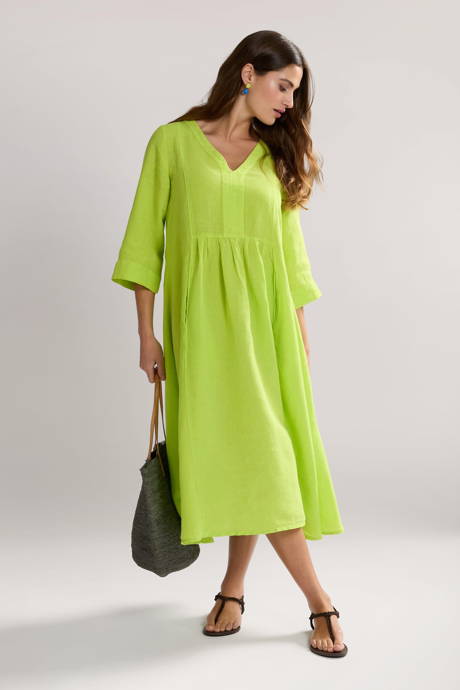 A woman in a flared silhouette, lime green, knee-length Organza Linen Pleat Front Dress with long sleeves holds a dark woven bag. She is wearing sandals and has long, wavy hair.