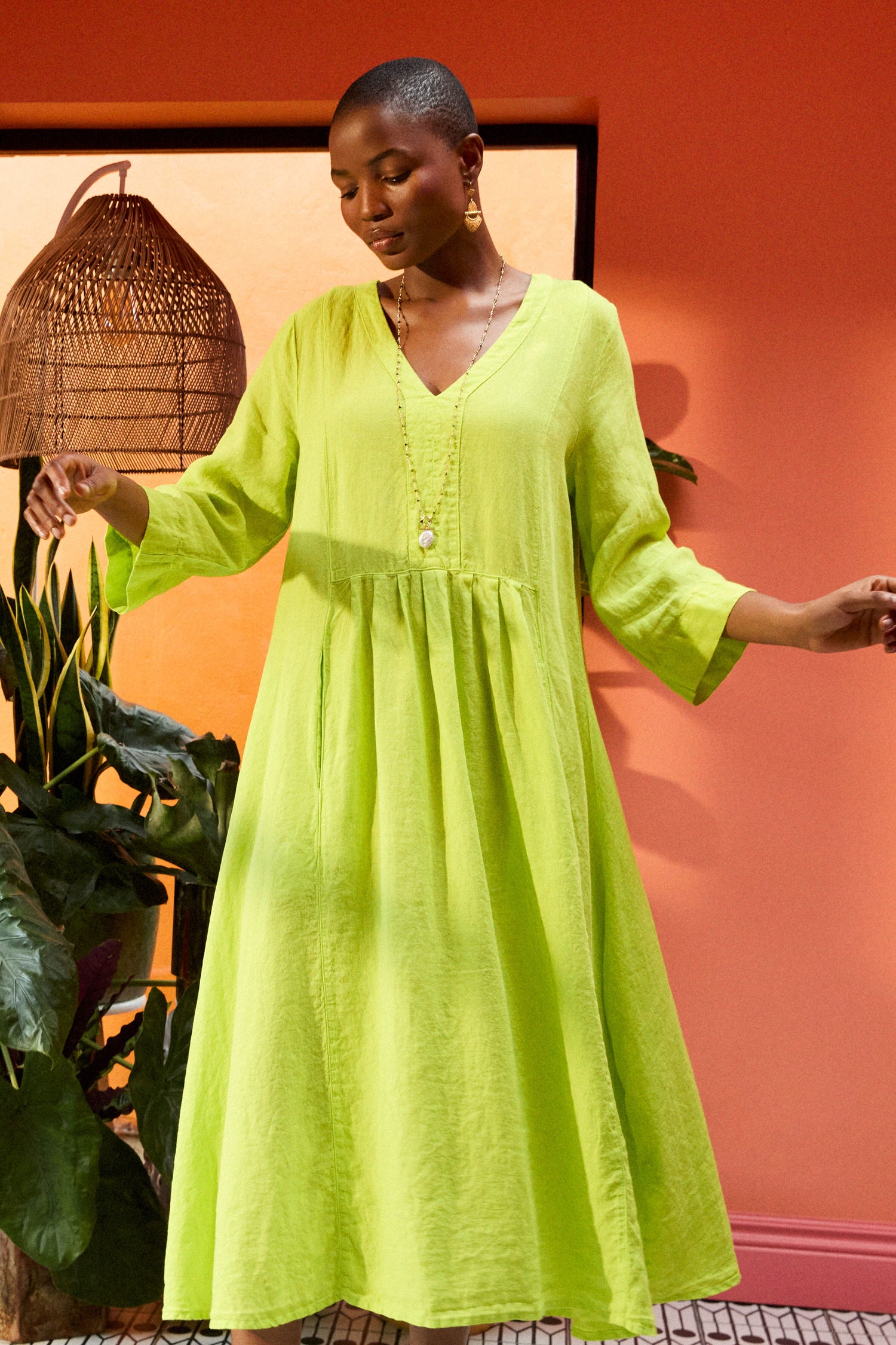 A person wearing an Organza Linen Pleat Front Dress stands in front of a peach-colored wall and potted plants. They have short hair and are accessorized with a long necklace and earrings.