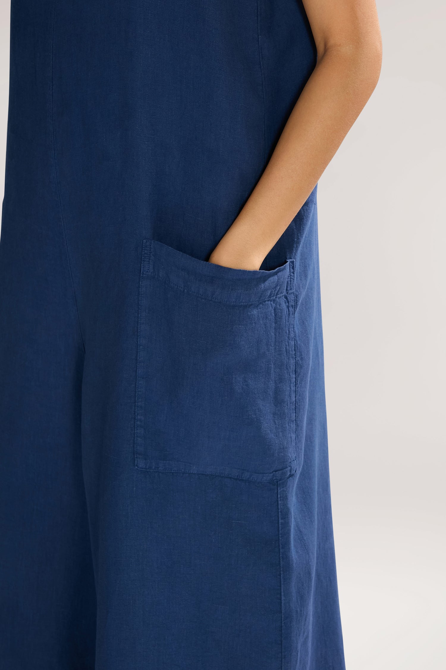 A close-up of a person wearing a blue Organza Linen Jumpsuit with a square neck, their hand tucked into the large front pocket.