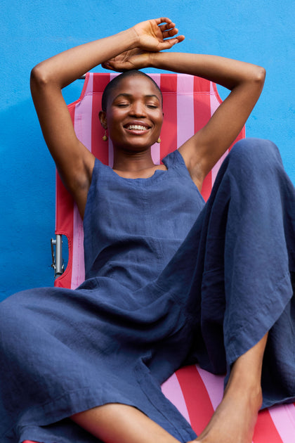 A person in a blue Organza Linen Jumpsuit reclines on a red and pink-striped chair, smiling with arms raised above their head, against a blue background. The wide leg design and oversized side pockets add both comfort and style to their relaxed pose.