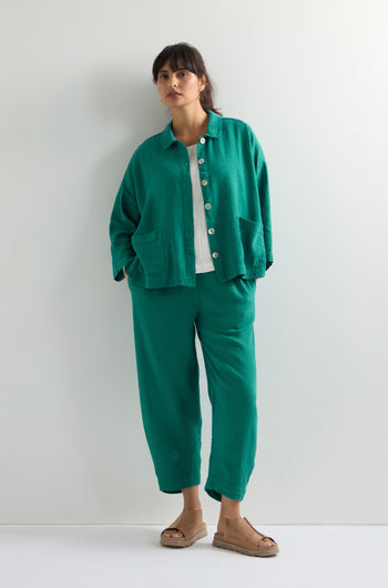 Against a white wall, someone wears the Twisted Linen Boxy Jacket in an oversized green style with matching pants, a crisp white shirt, and sandals.