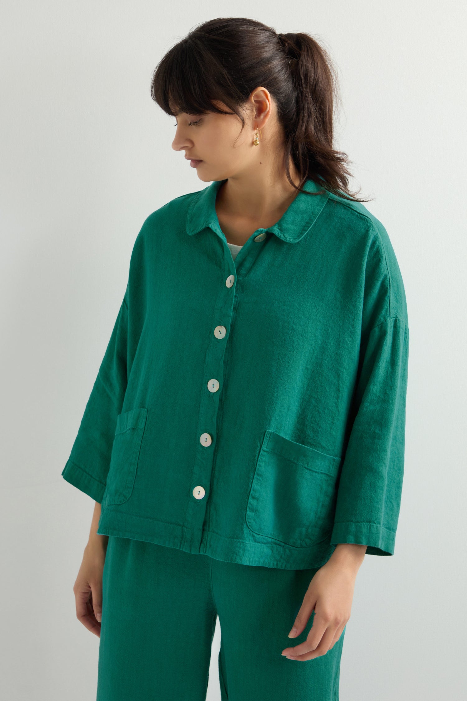 A person in a Twisted Linen Boxy Jacket stands against a plain background, looking down. Their green button-up shirt and matching pants resemble an oversized fit with the ease of a breathable linen blend, enhancing the relaxed vibe.