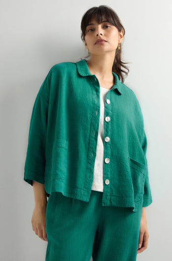 A person wears the Twisted Linen Boxy Jacket, a loose green button-up with an oversized fit over a white top, paired with green breathable linen blend pants, standing against a plain background.