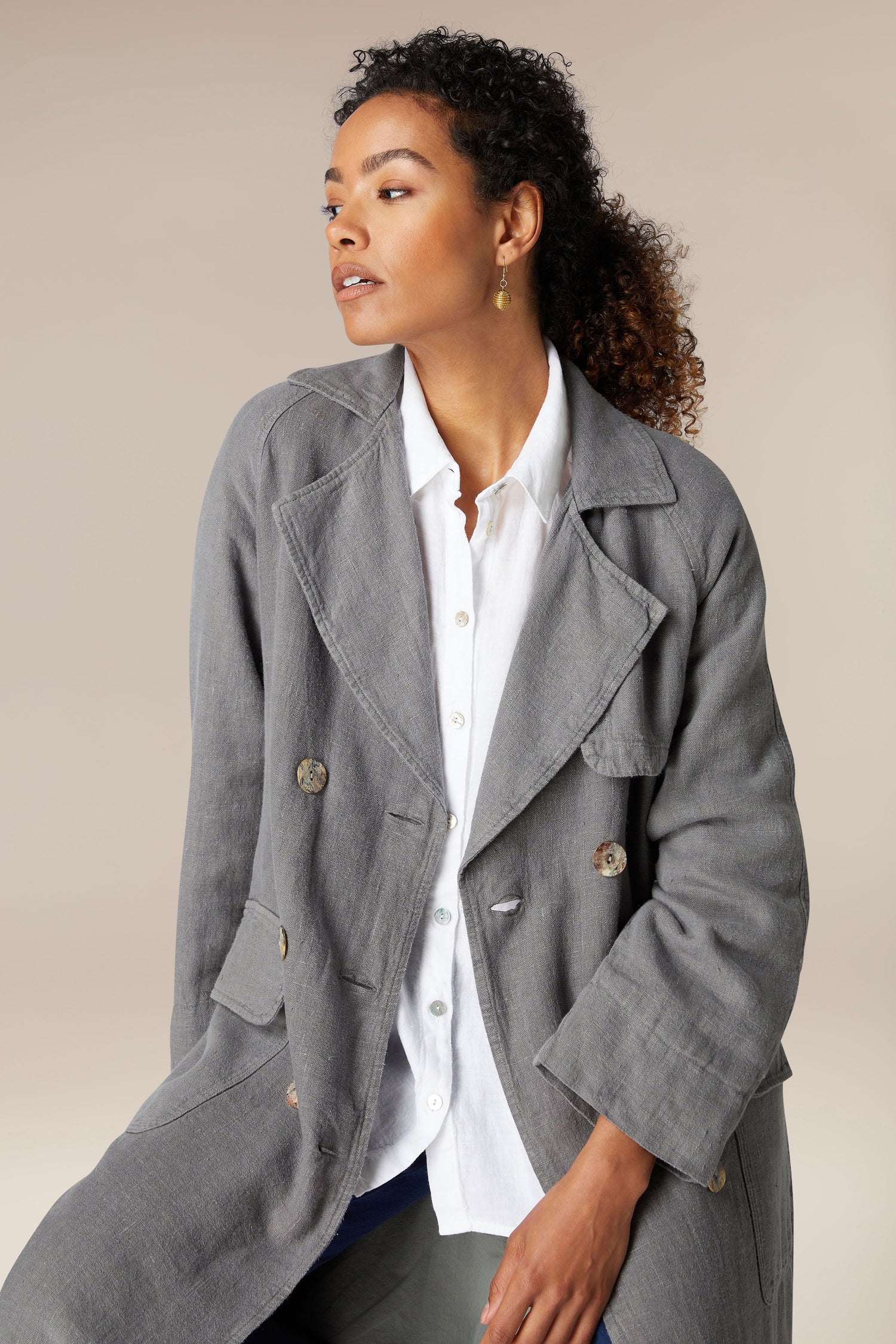 A woman wearing a stylish gray linen trench coat over a white button-up shirt, posing against a neutral background.