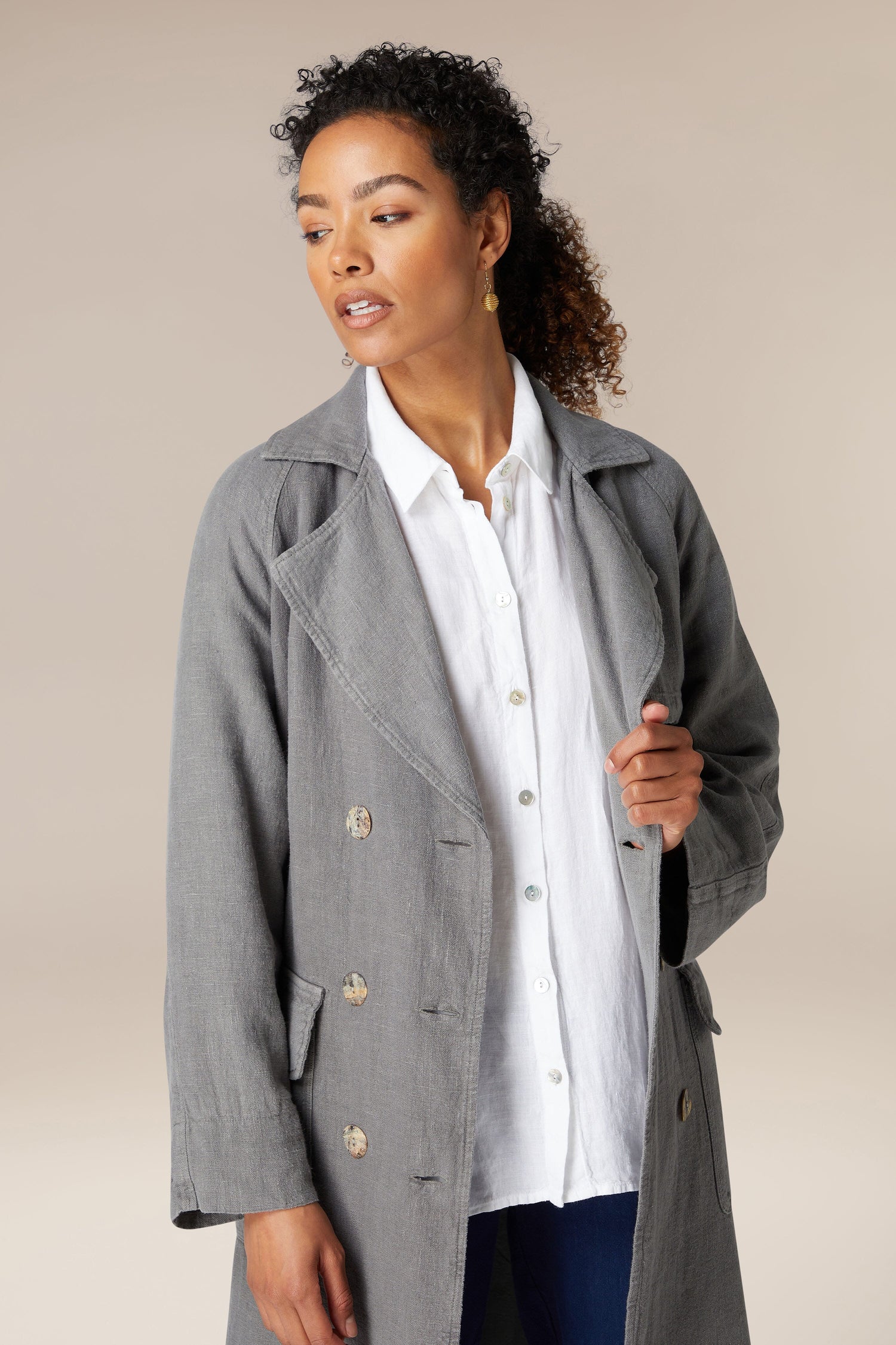 A woman in a stylish gray Linen Trench Coat and white shirt, looking to the side with a slight smile, against a neutral background.