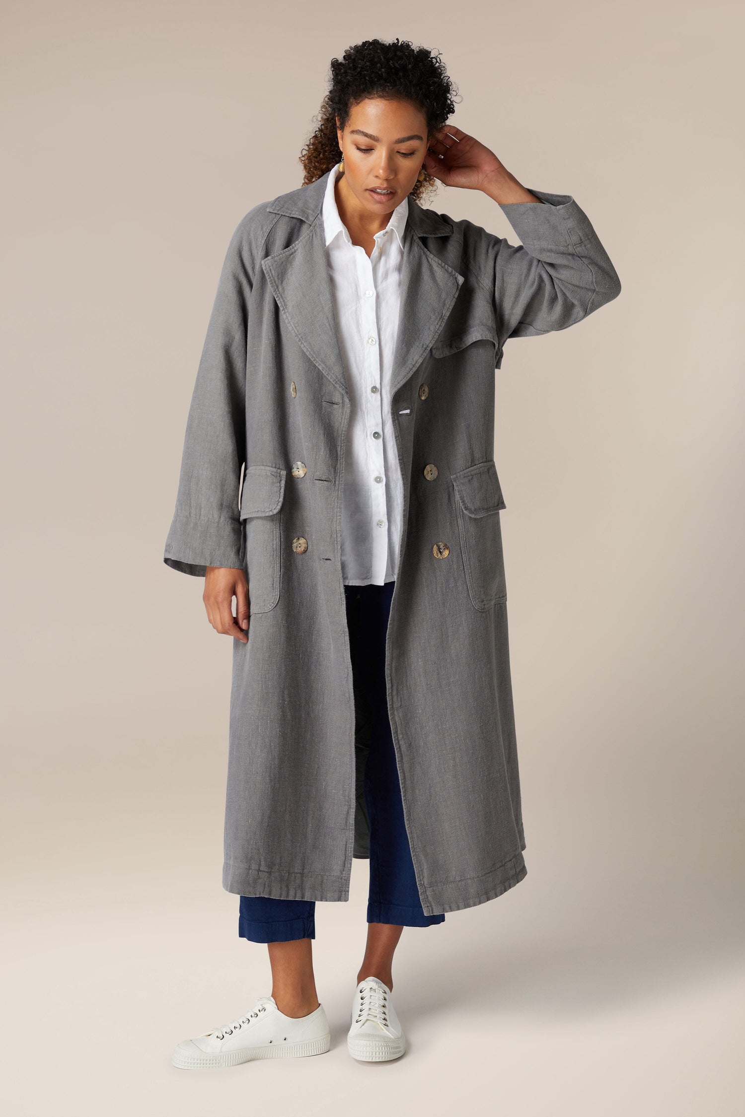 A woman in a stylish gray Everlane Linen Trench Coat, white shirt, blue jeans, and white sneakers poses in a neutral studio setting.