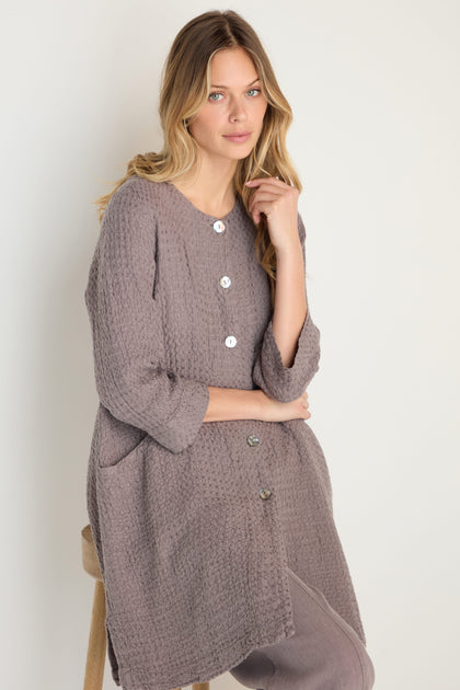 A woman with long blonde hair wears the Waffle Linen Long Jacket and matching pants, true wardrobe essentials, while sitting on a stool against a plain background.