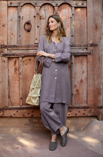 A woman wearing a purple Waffle Linen Long Jacket and pants stands by a rustic wooden door, holding a woven bag and looking to the side.