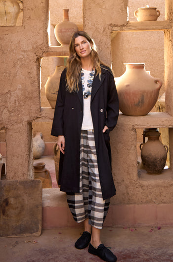 A woman stands indoors in a rustic setting, wearing the Twisted Linen Relaxed Coat, a white printed shirt, and black-and-white plaid pants, surrounded by large clay pots on shelves.