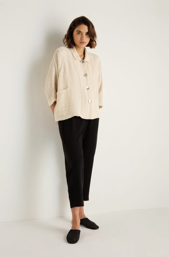 A person stands against a white wall wearing a cream textured button-up jacket, Twisted Linen Slim Trouser in black, and black slip-on shoes.